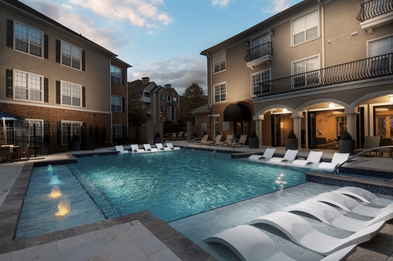 Outdoor pool surrounded by apartment buildings with lounge chairs partially submerged in the water. Tables and chairs are positioned on the pool deck.