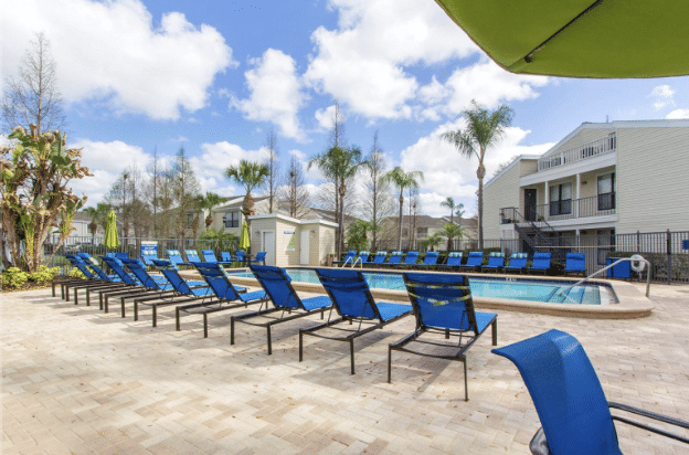 Outdoor swimming pool surrounded by rows of blue lounge chairs, palm trees, and a few buildings under a partly cloudy sky.