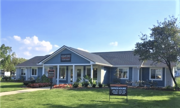 Exterior view of a one-story office building with a sign reading "Ashford Office Center." It has a landscaped yard and a sign indicating office hours.