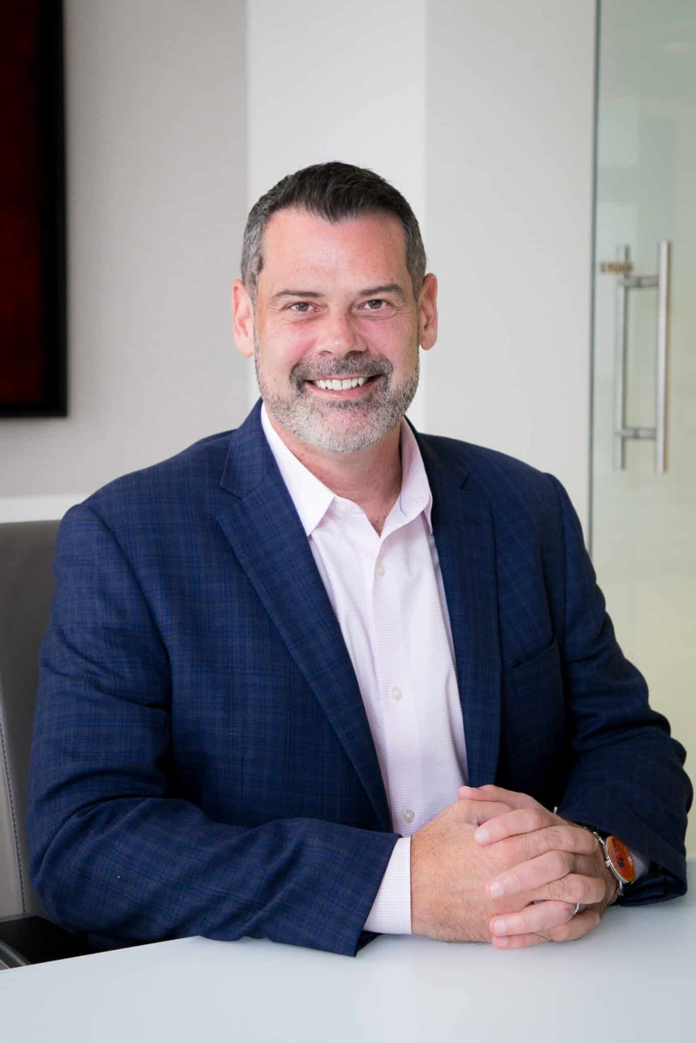 A man with a beard in a blue suit and white shirt sits smiling at a table in a modern office setting.