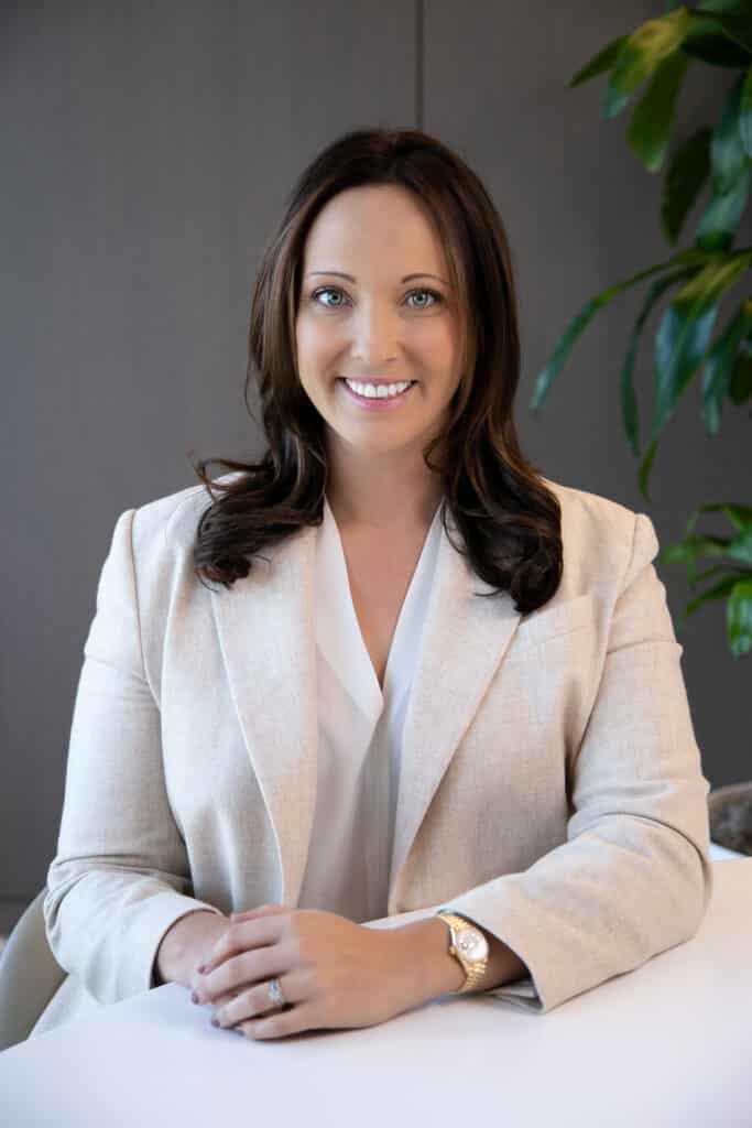 A person with long dark hair sits at a table, wearing a light-colored blazer and white blouse, with hands folded and a plant in the background.