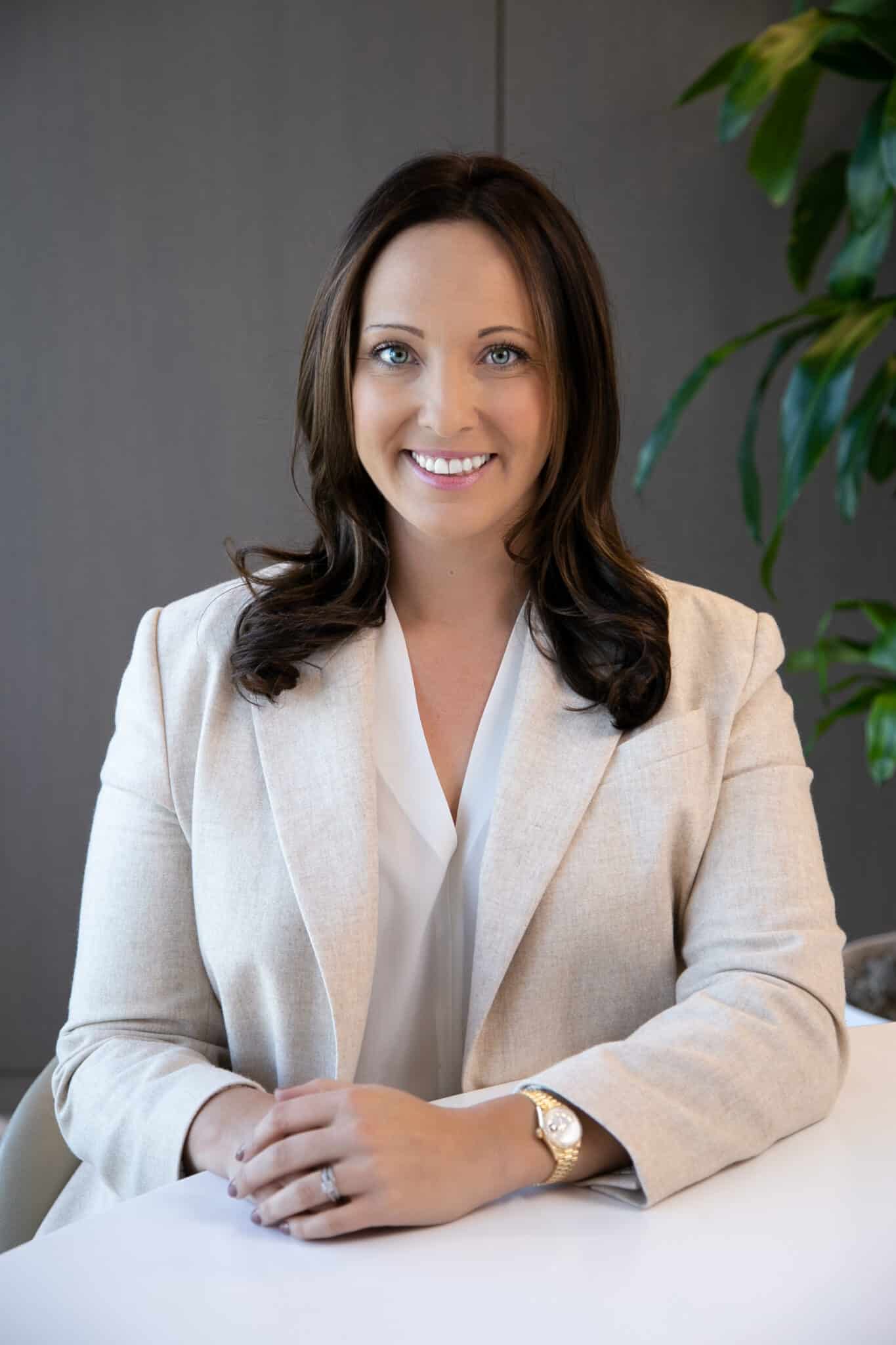 A person with long dark hair sits at a table, wearing a light-colored blazer and white blouse, with hands folded and a plant in the background.