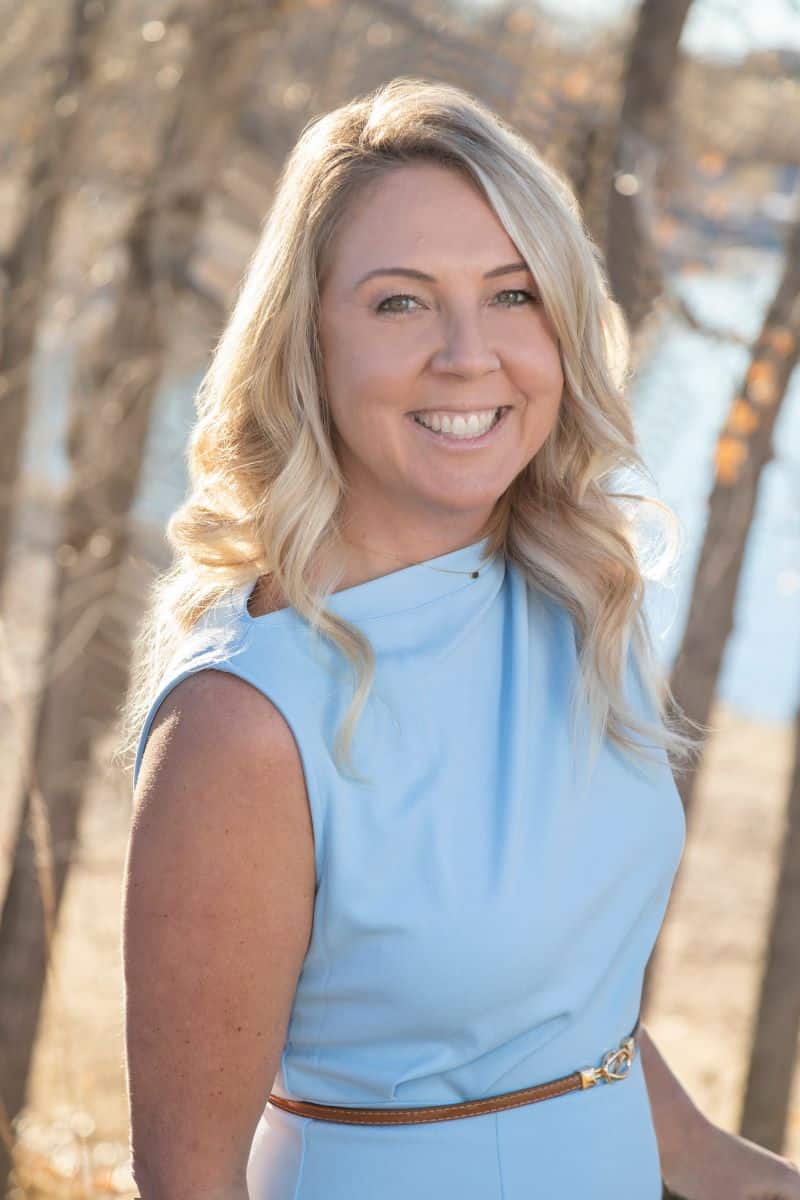 A woman with long blonde hair and a blue sleeveless dress smiles outdoors with trees and water in the background.