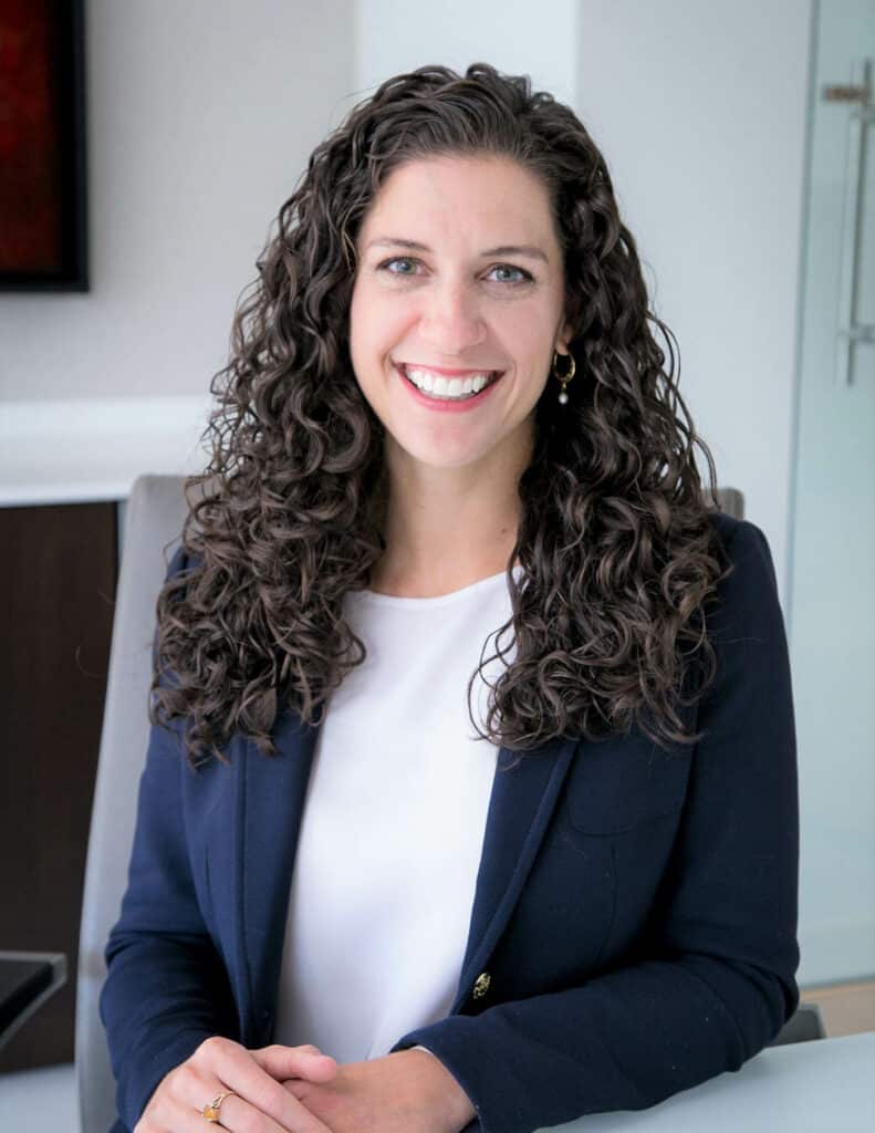 A woman with long curly hair, dressed in a navy blazer and white blouse, is smiling while seated at a desk in an office setting.