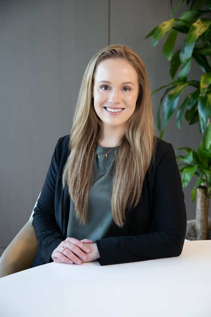 A woman with long blonde hair, wearing a black blazer and a green top, is seated at a white table, smiling with her hands folded in front of her. A green plant is visible in the background.