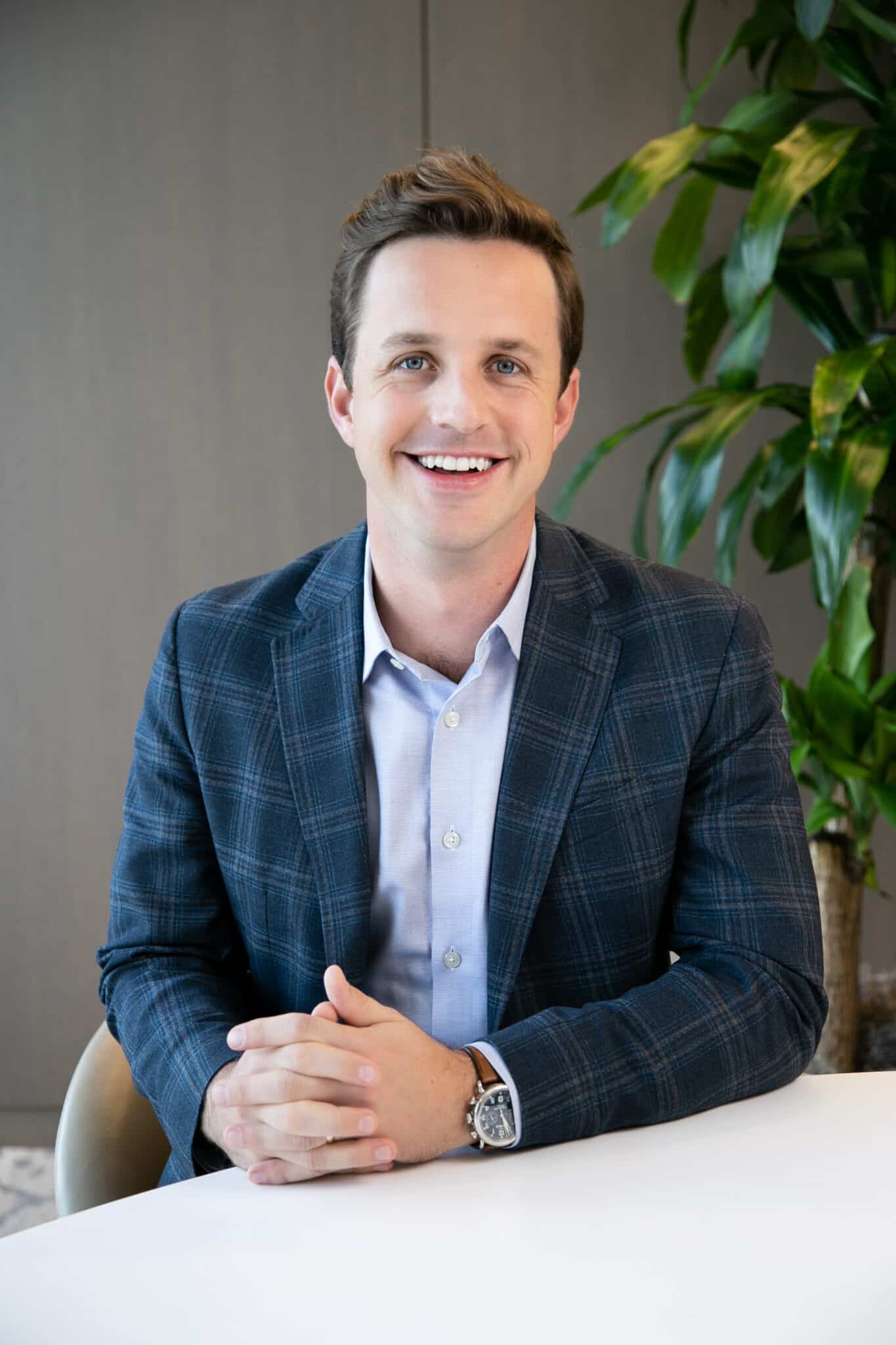 A man in a checkered blazer and light blue shirt sits at a white table, smiling. A plant is visible in the background.