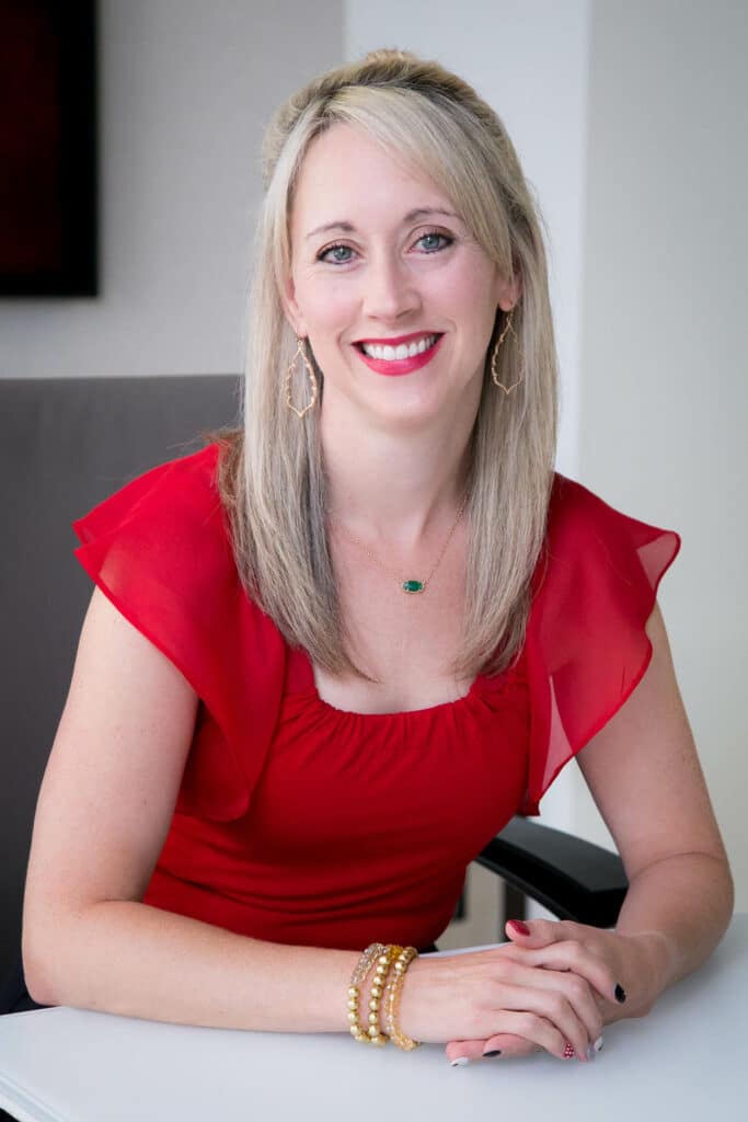 A person with long blonde hair wearing a red top, sitting at a table and smiling.