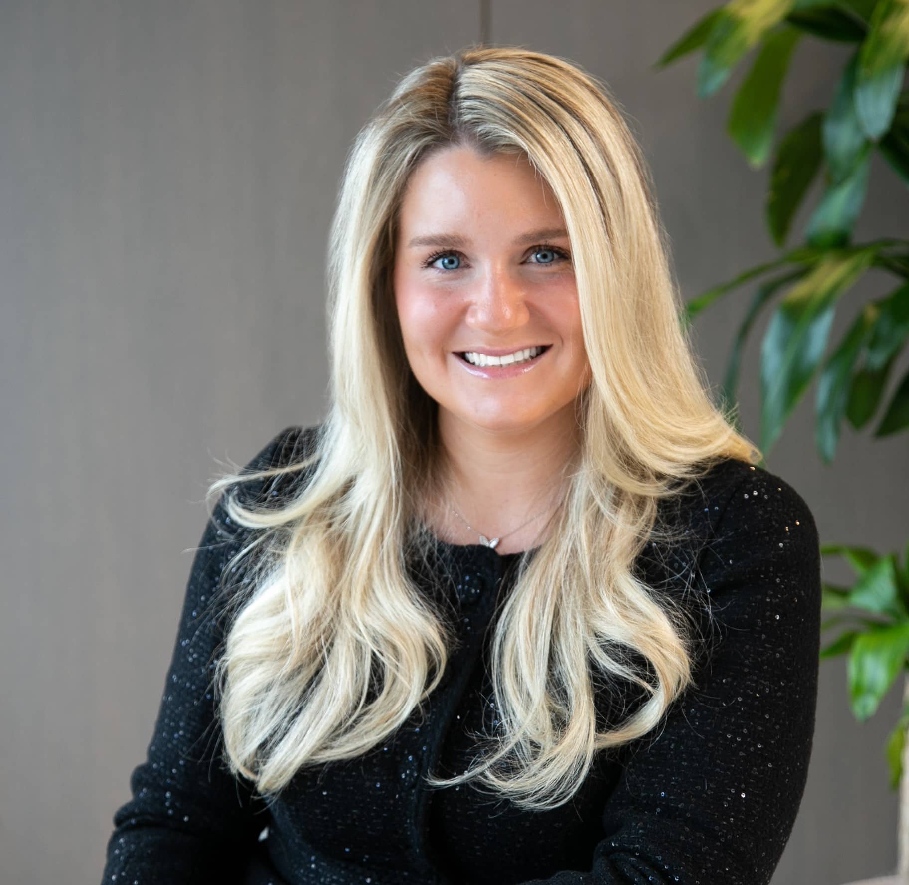 A person with long blonde hair wearing a black top, seated indoors with a plant visible in the background.