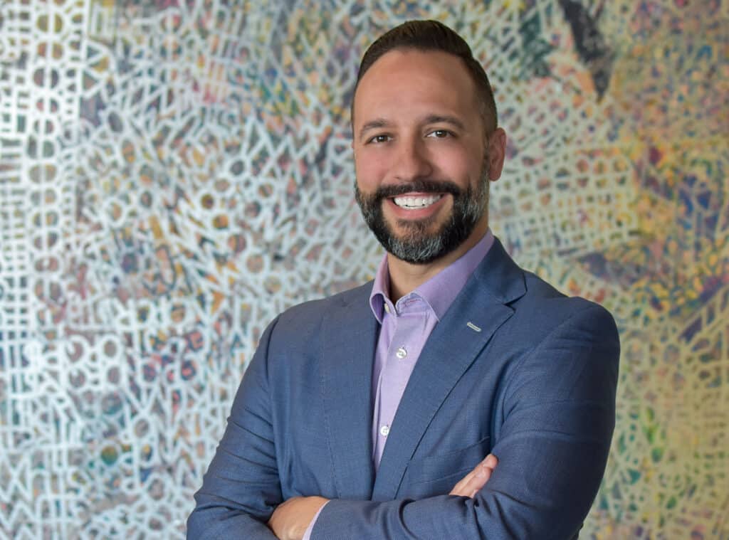 A man with a beard, wearing a blue suit and a light purple shirt, stands with his arms crossed in front of a colorful, abstract background. He is smiling.