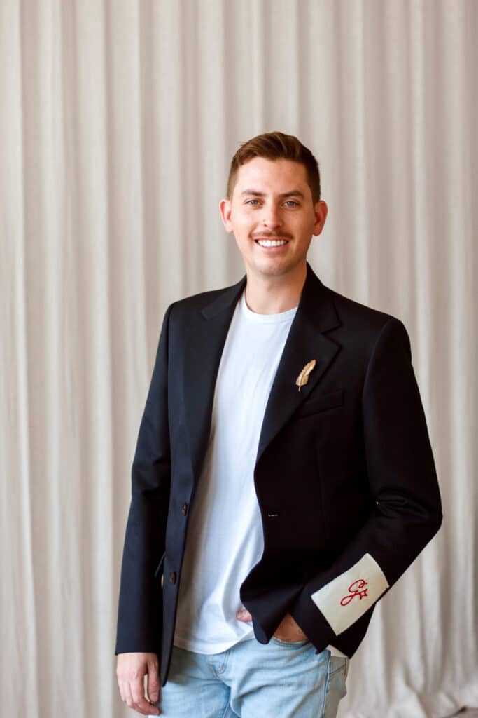 A man with short hair and a mustache is wearing a black blazer, white t-shirt, and jeans. He stands in front of a neutral curtain background, smiling and holding a pen.