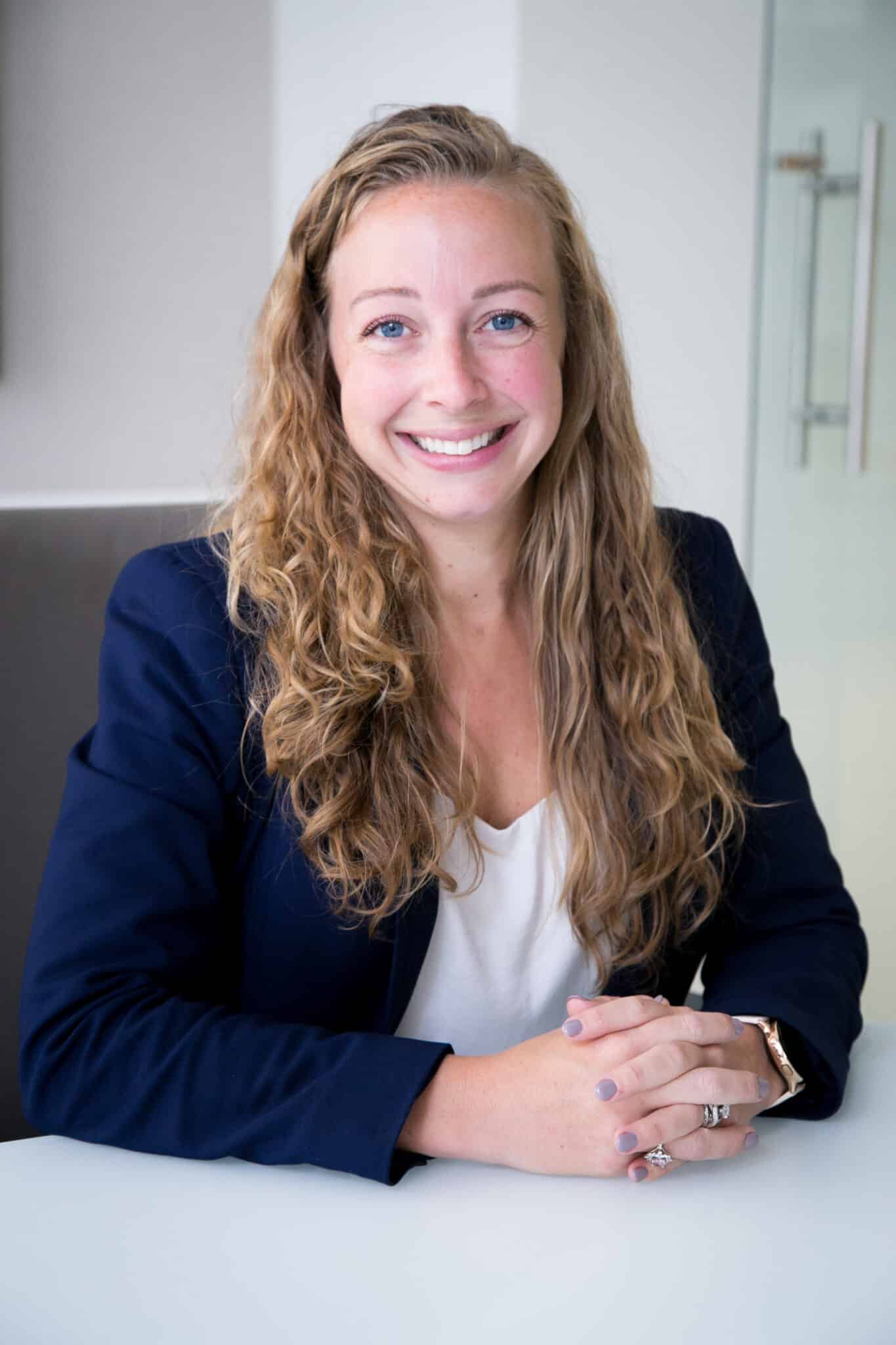 A woman with long curly hair, wearing a navy blazer and white shirt, is seated at a table, smiling, with her hands clasped in front of her.