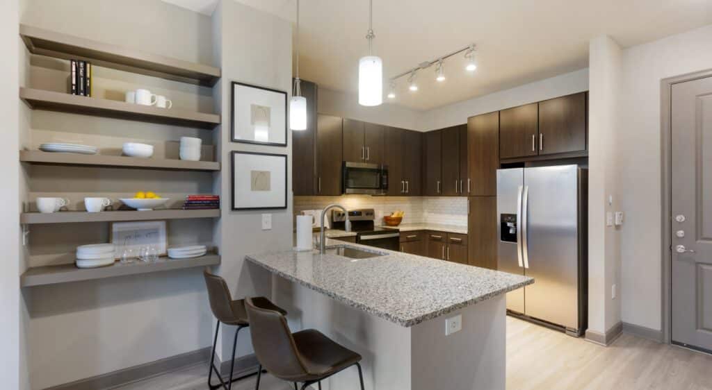 Modern kitchen with brown cabinets, stainless steel appliances, and a granite countertop island with two barstools. Open shelving displays dishware and framed pictures adorn the wall. .