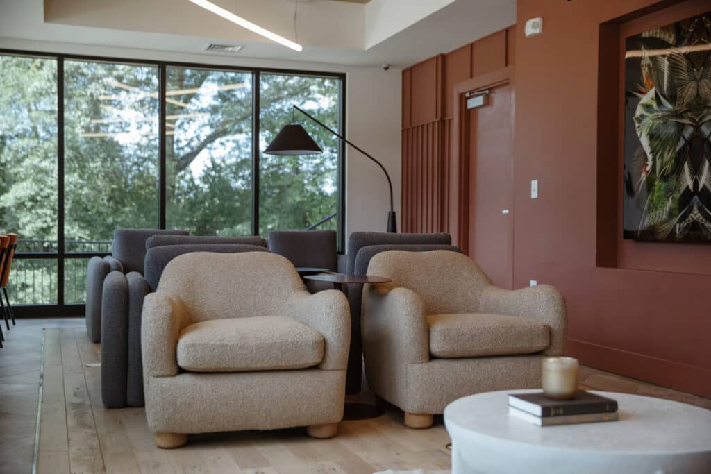 The image shows a cozy lounge area with beige armchairs, a dark lamp, a large window with a view of trees, and a modern interior design. A book and a candle are on a nearby coffee table.