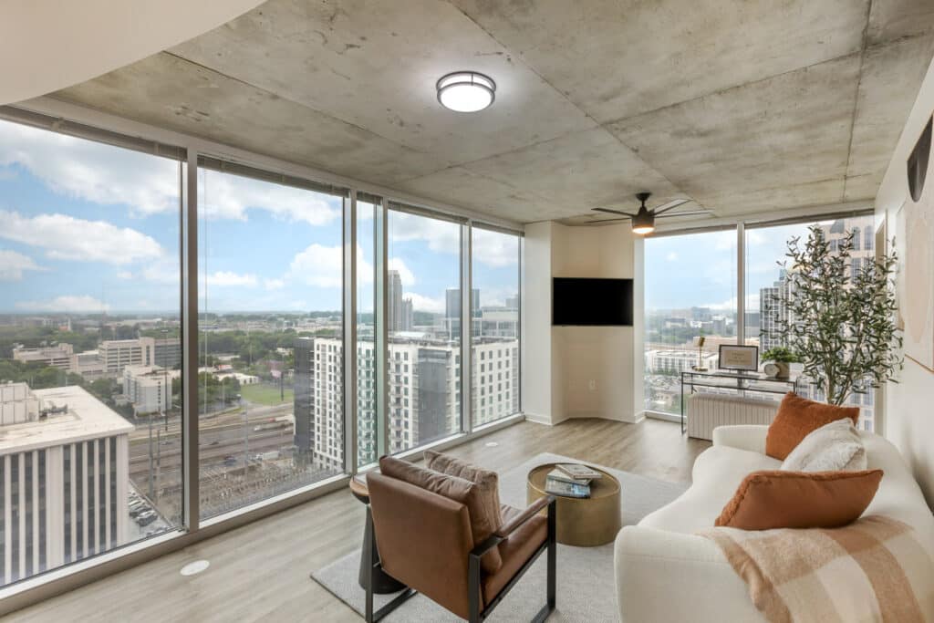 A modern living room with floor-to-ceiling windows showcasing a cityscape. The space features a white sofa, brown armchair, a TV mounted on the wall, and a ceiling fan. A small table stands between the seats.