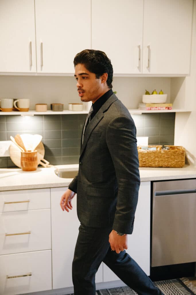 A man in a dark suit walks through a modern kitchen with white cabinets, a backsplash, and various kitchen items on the counter.