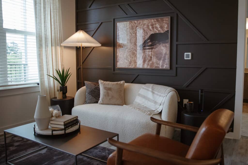 A modern living room with a white sofa, brown leather chair, lamp, and coffee table with books and decor items. A framed picture is mounted on the dark accent wall behind the sofa.