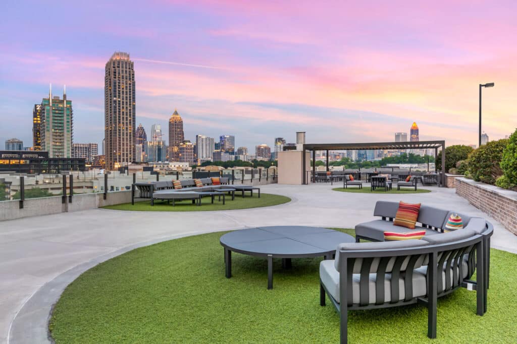 Rooftop lounge with modern seating, artificial grass areas, and city skyline at sunset in the background.