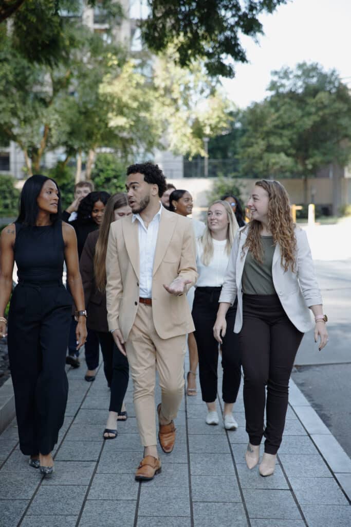 A group of adults in professional attire are walking and talking on a paved pathway outdoors, surrounded by trees.