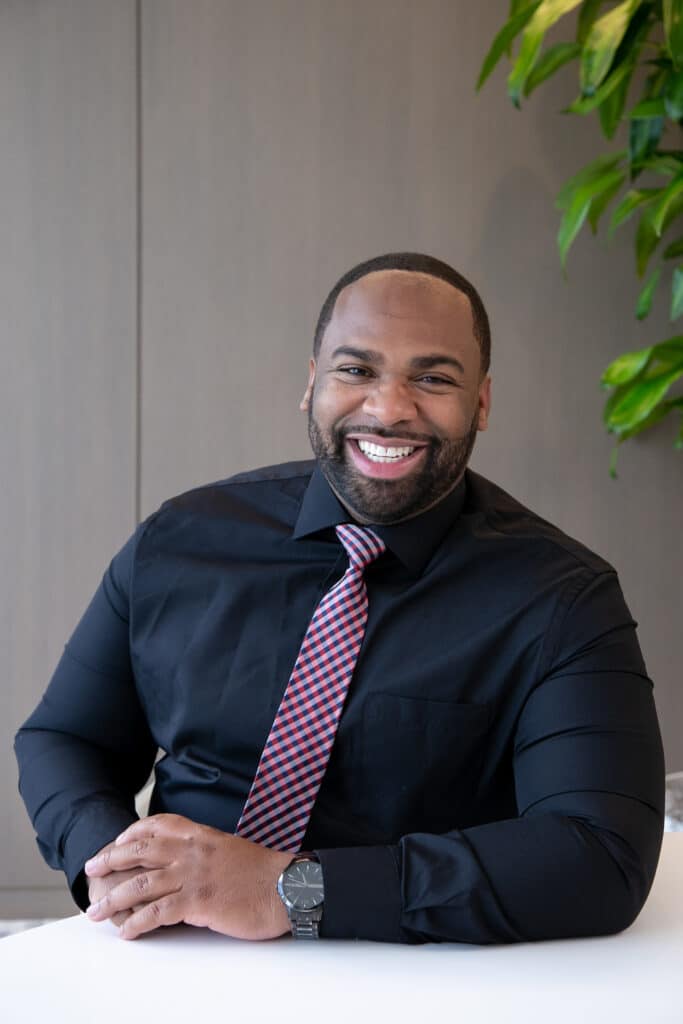 A man wearing a black dress shirt and a red-striped tie is smiling, seated at a table with a plant in the background.