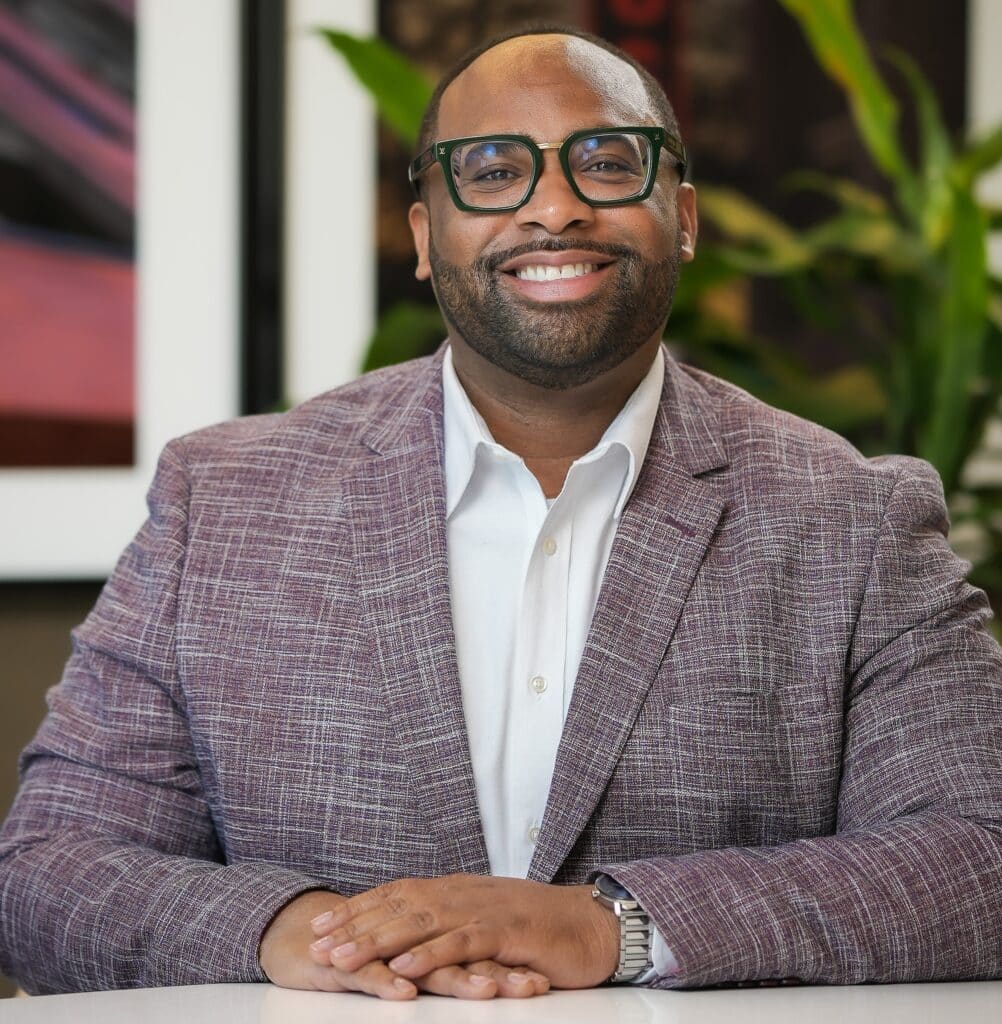 A man wearing glasses and a checked blazer sits at a table with his hands folded, smiling at the camera. A plant and framed artwork are visible in the background.