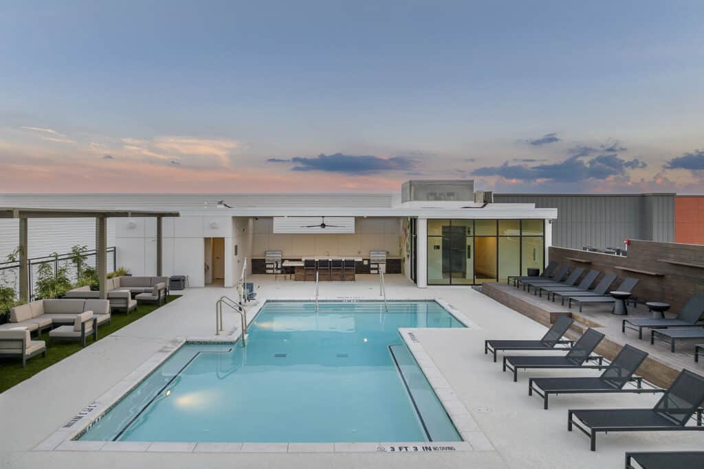 A rooftop pool features lounge chairs, seating areas, and a nearby building with large windows, set against a sunset sky.