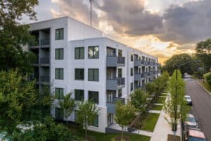 A modern, four-story apartment building at sunset, characterized by white and gray exterior walls, large windows, and small balconies. The street features parked cars and a row of young trees.