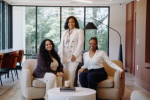 Three women are posing in a modern, well-lit office space. Two are seated on armchairs and one is standing behind them. Large windows and minimalistic decor are visible in the background.