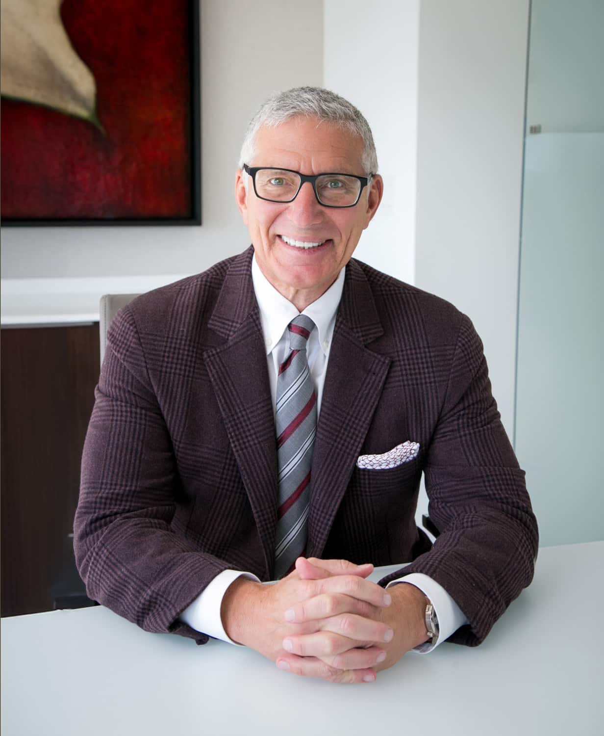 A man with glasses and gray hair, wearing a plaid suit jacket and striped tie, sits at a desk with his hands clasped.