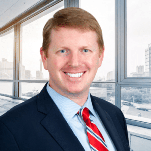 Man in a suit and red striped tie smiling, standing in front of large office windows with a cityscape in the background.
