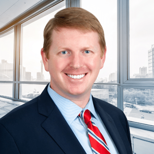 Man in a suit and red striped tie smiling, standing in front of large office windows with a cityscape in the background.